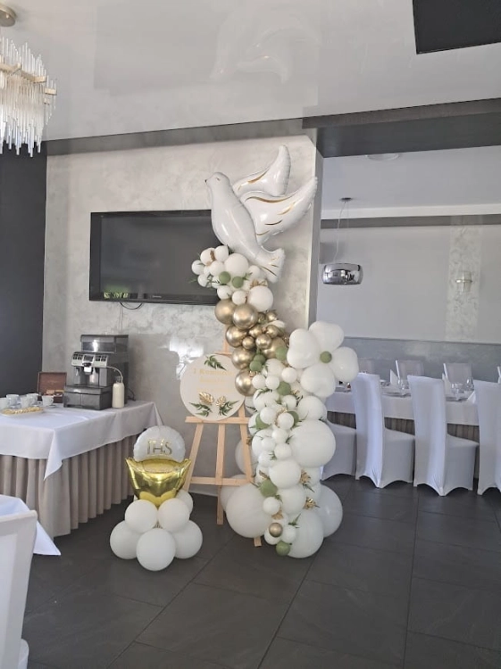 Ścianka do zdjęć na komunię z gołębiem, elegancka dekoracja sali na sztaludze, balony białe i złote z zielenią. Duży balonowy gołąb  z mini słupem.