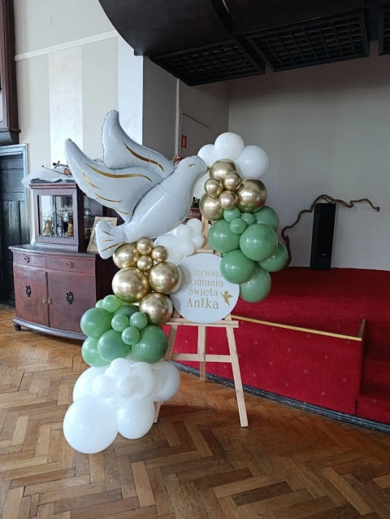 Ścianka do zdjęć na komunię z gołębiem, elegancka dekoracja sali na sztaludze, balony białe i złote z zielenią.